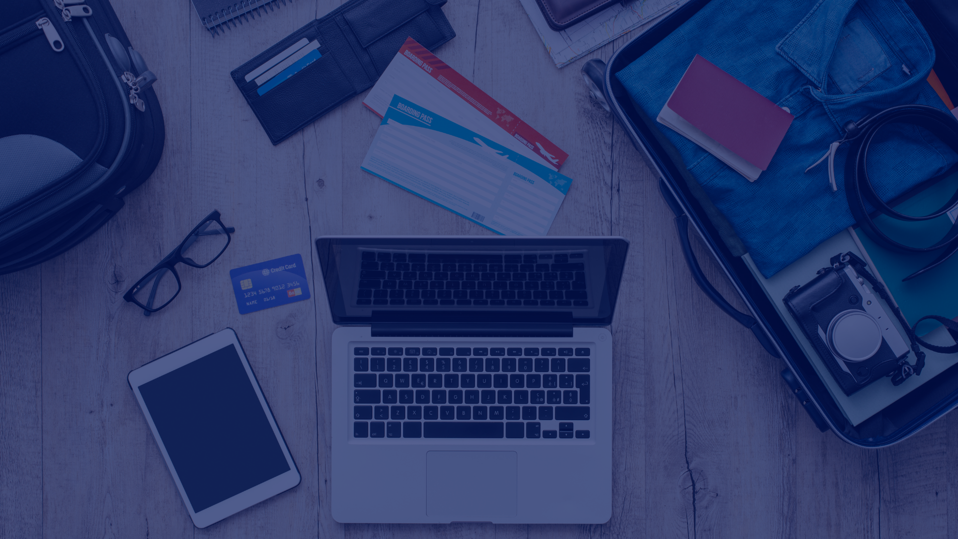 A top-down shot reveals a laptop, tablet, and smartphone alongside boarding passes, wallets, glasses, and a packed suitcase on a light wooden surface. The arrangement conveys a sense of preparation for a journey, blending technology with traditional travel items. The open laptop and other devices suggests planning and connectivity on the go. The photo's bright lighting and organized composition lends itself well to concepts such as travel planning, digital nomadism, technology integration, and vacation preparation. Its neutral tones and clear imagery make it suitable for use in travel blogs, technology websites, or marketing materials for travel agencies.