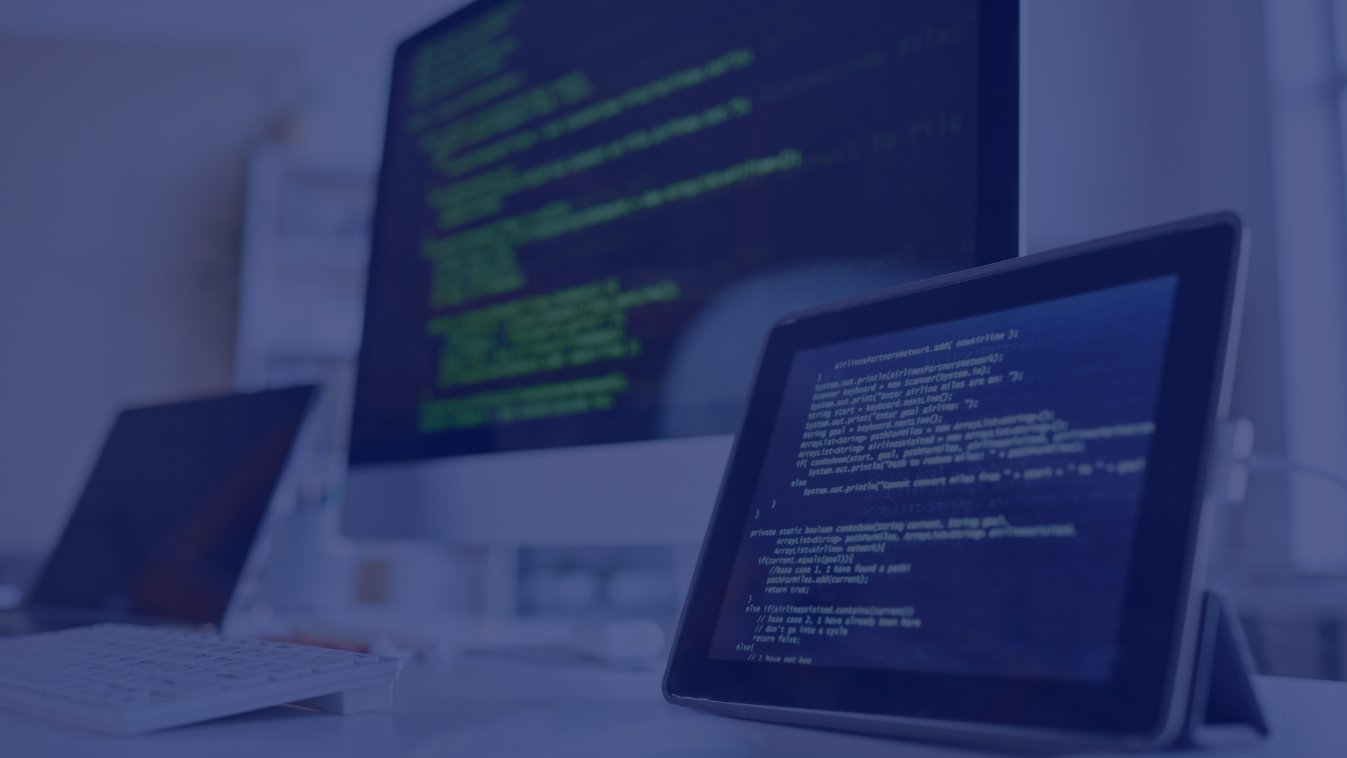 This photo showcases a brightly lit office setting, dominated by a computer and tablet displaying lines of code. A partially visible laptop and keyboard suggest a workspace focused on technology. The setup, against a blurred backdrop, evokes a modern and professional atmosphere. This image is ideal for projects centered on programming, software development, or digital workspaces, embodying themes of innovation, precision, and technological advancement.