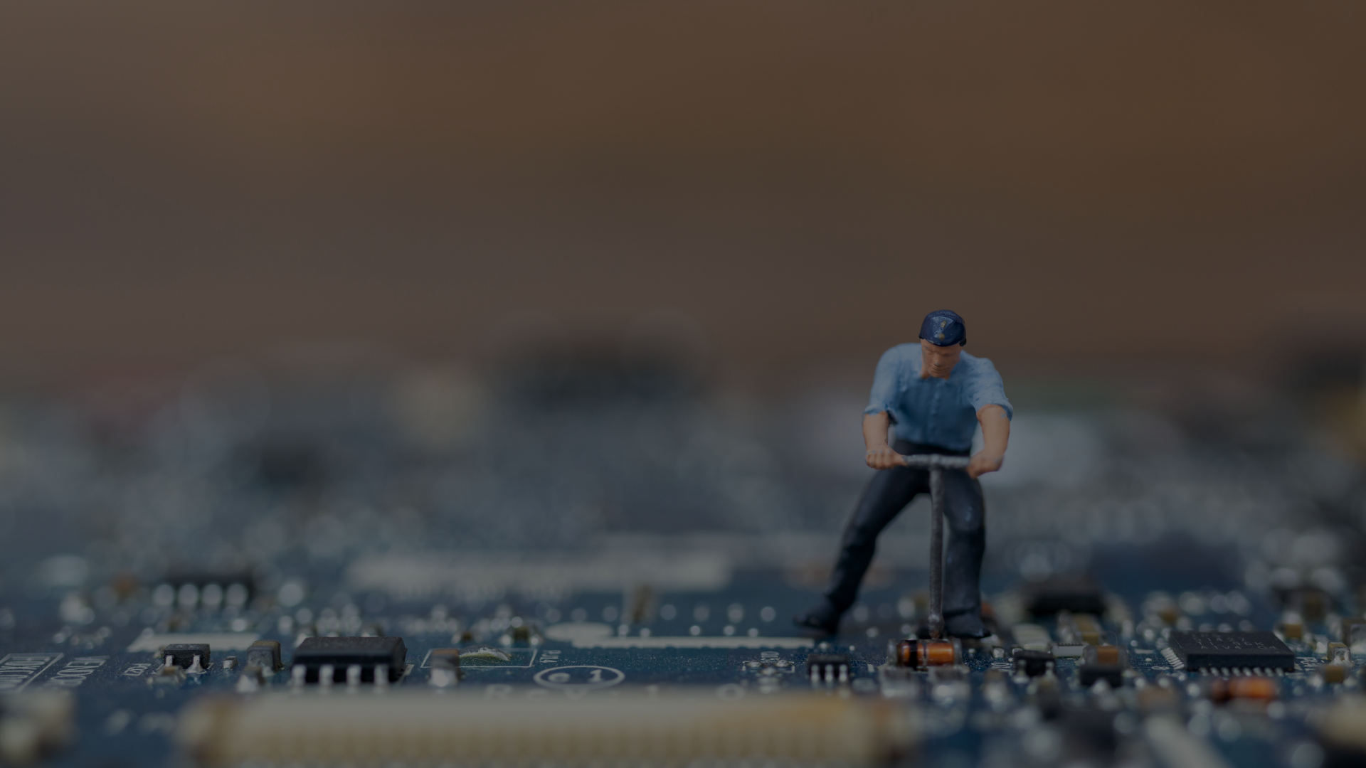 A tiny figurine of a man with short brown hair, wearing a blue short-sleeved shirt, dark trousers and a blue baseball cap, uses a tool on a blue circuit board. He is a product engineer. The image evokes the idea of fixing or inspecting computer or technology components. The background is out of focus. The mood is serious and analytical. This photo could be used for illustrative purposes in business or educational materials about technology, engineering, or innovation.